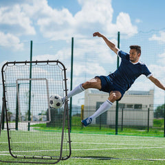Ein Fußballspieler führt einen dynamischen Seitfallzieher vor dem großen Rebounder auf einem Sportplatz aus.