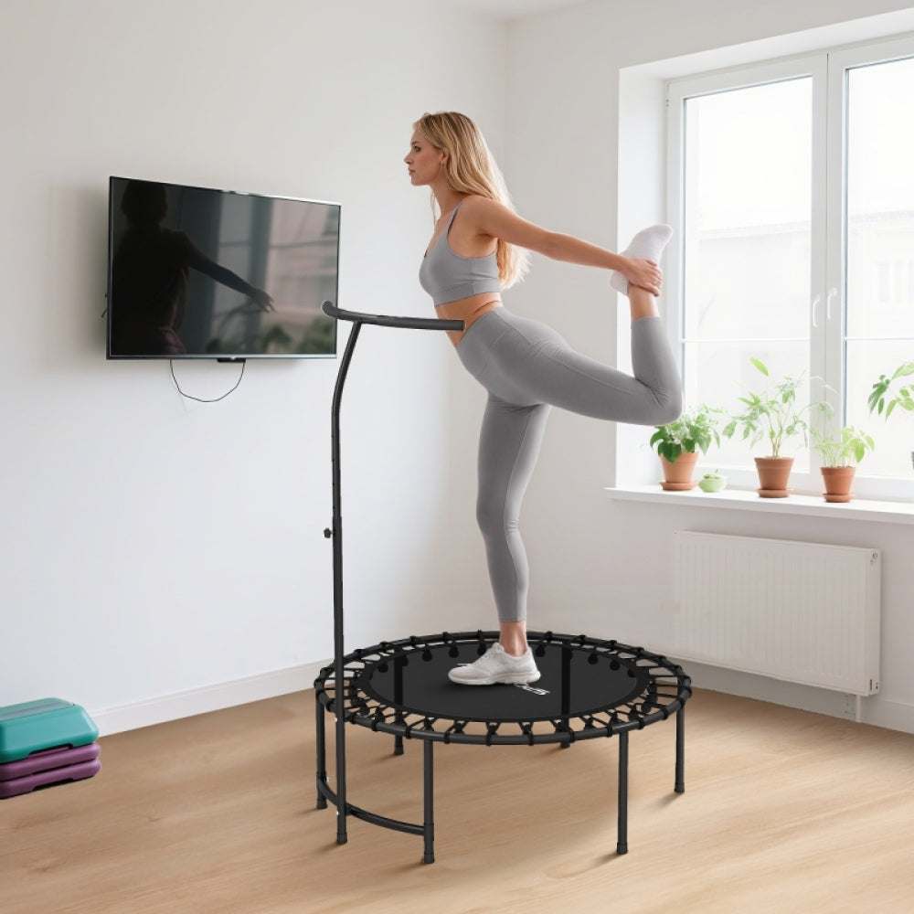 Frau dehnt ihr Bein auf dem Trampolin und hält sich am hohen T-Griff fest für Balance.